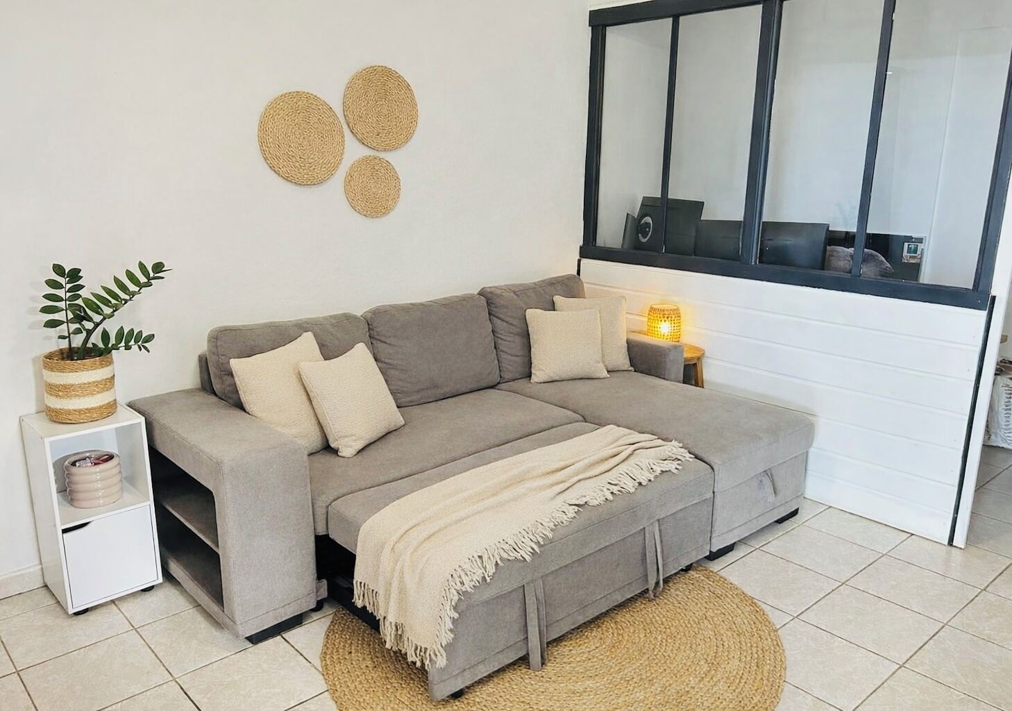Un salon moderne avec un canapé gris, une petite table d’appoint avec une lampe, un meuble blanc et une grande fenêtre.