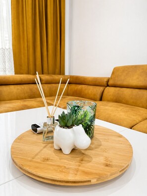 A round wooden table with a white surface, a white ceramic planter with a succulent, a glass container with green leaves, and a white electronic device. - Pami Apartments (Nea Peramos)