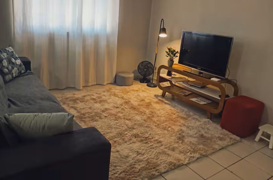 Un salon avec un canapé, un téléviseur posé sur un meuble en bois, un petit ventilateur et un pouf rouge.