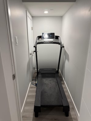 Un pasillo estrecho con una cinta de correr en el centro, una puerta a la izquierda y una lámpara en el techo.