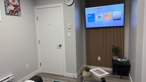 A modern living room with a flat-screen TV displaying a calendar, a white door, a wall clock, a small table with a plant, and a decorative painting. - New 1BR Suite in Aspen Ridge, Saskatoon for a home away from home experience. (Saskatoon)