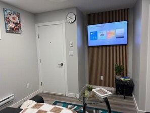 Uma sala de estar moderna com uma televisão de tela plana exibindo um calendário, uma porta branca, um relógio de parede, uma pequena mesa com uma planta e uma pintura decorativa.
