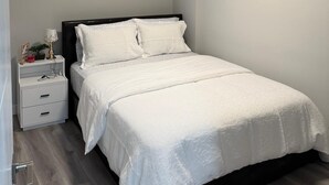 A bedroom with a bed, a nightstand, a window with blinds, a ceiling light, and a wall-mounted picture. - New 1BR Suite in Aspen Ridge, Saskatoon for a home away from home experience. (Saskatoon)