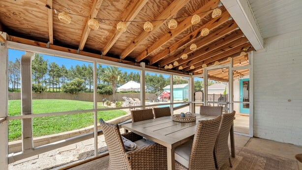 Een overdekt terras met een tafel en rieten stoelen, grote ramen en een houten plafond.