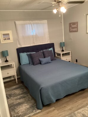 A bedroom with a bed, two bedside tables, a nightstand, a ceiling fan, a window with a sheer curtain, and a wall sign.
