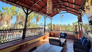 Eine Terrass mit Holztisch, Korbstühlen, einem hängenden Dekorationsobjekt und Blick auf Palmen und Gebäude.