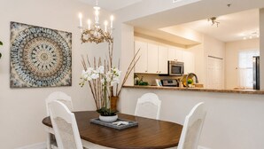 A dining area with a round table, white chairs, a chandelier, and a kitchen in the background. - Live the Naples Lifestyle.. Resort Amenities & Coastal Charm (Naples)