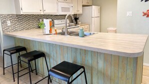 A kitchen with white cabinets, a microwave, a refrigerator, a sink, and two black bar stools. - High-Floor Saida Beachfront - Pool + Ocean View (South Padre Island)