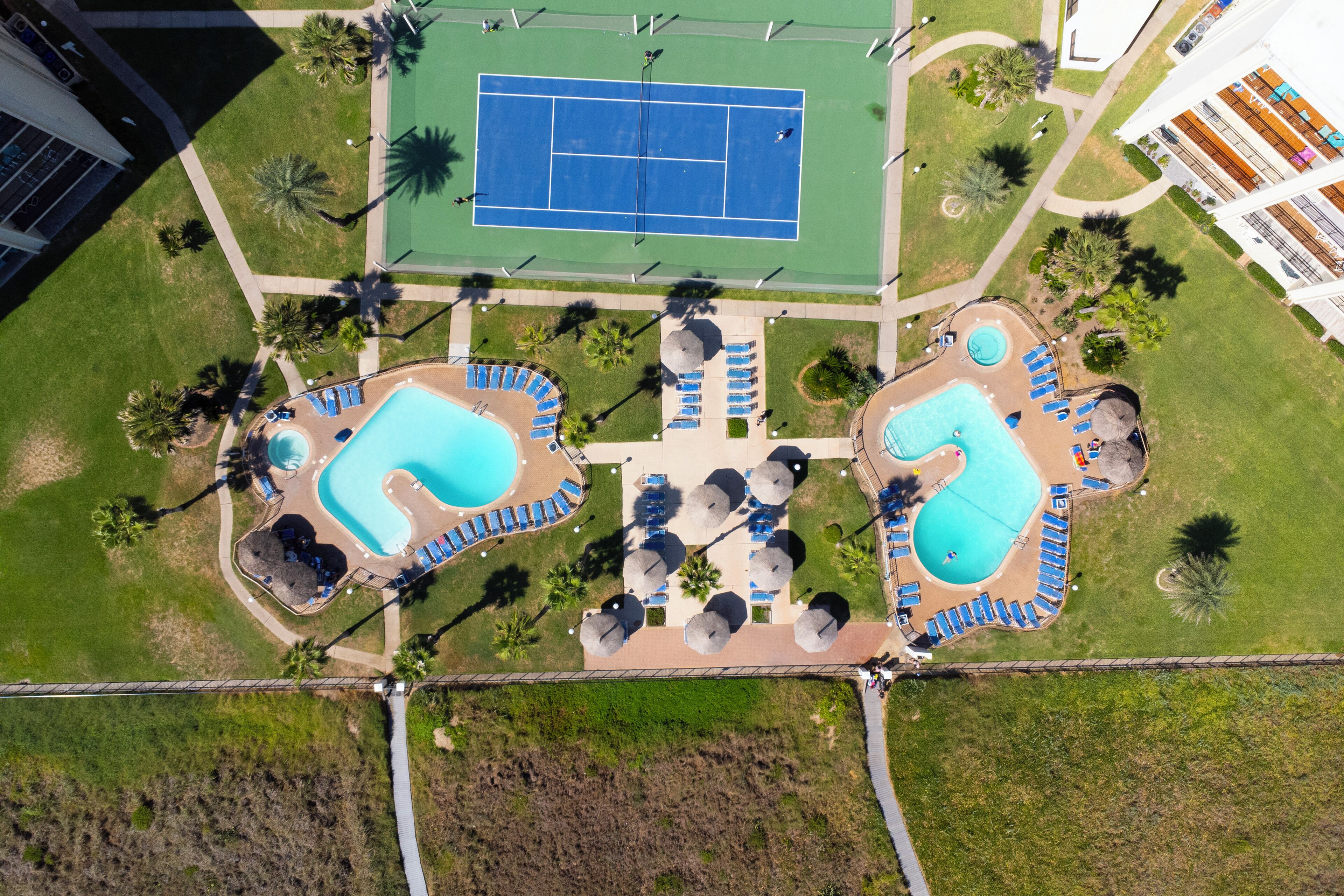 An aerial view of a resort with two swimming pools, tennis courts, and lounge areas.