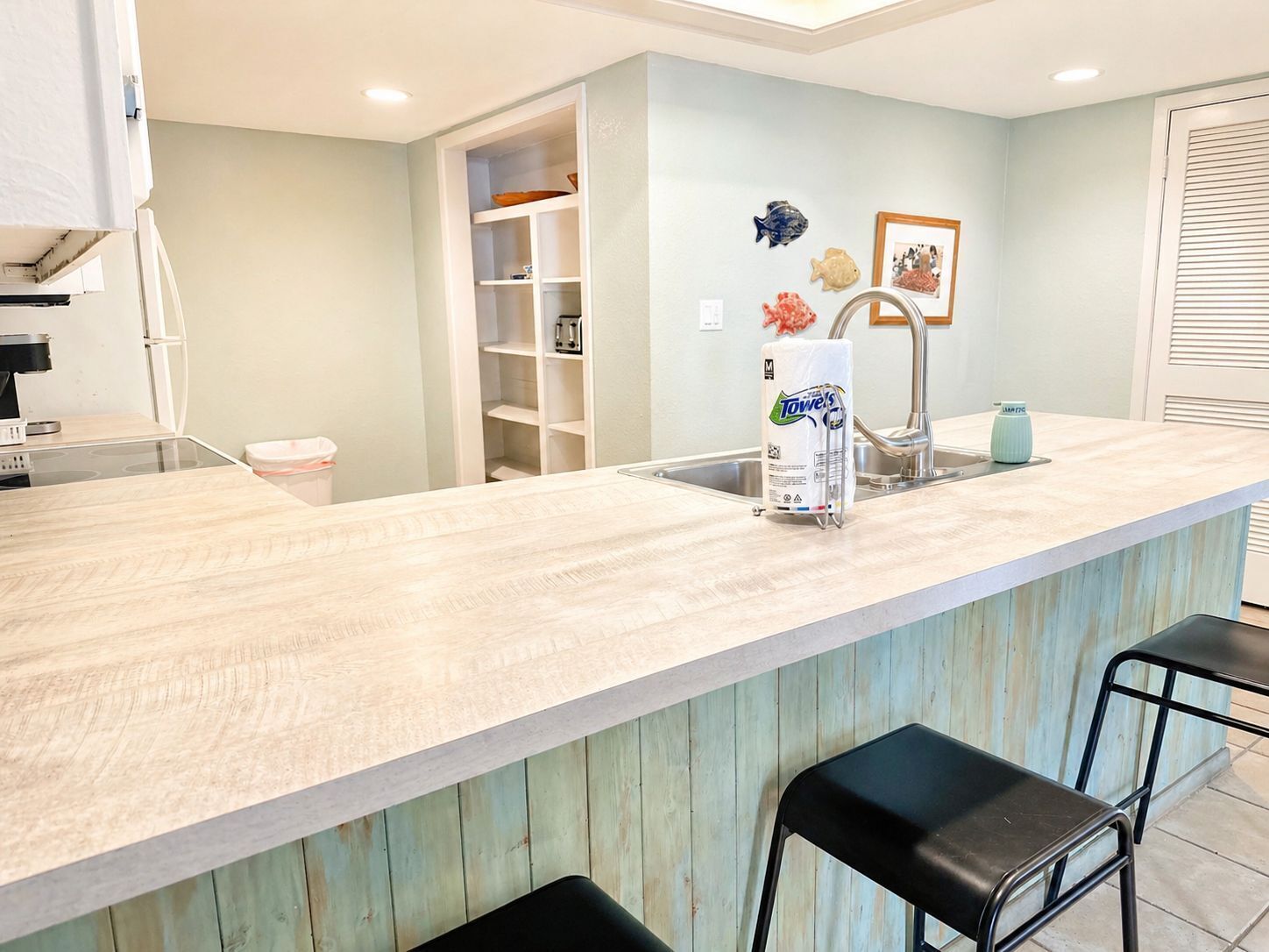 A kitchen with a light-colored countertop, a sink, and a towel dispenser. There are two black bar stools and a white cabinet with shelves.