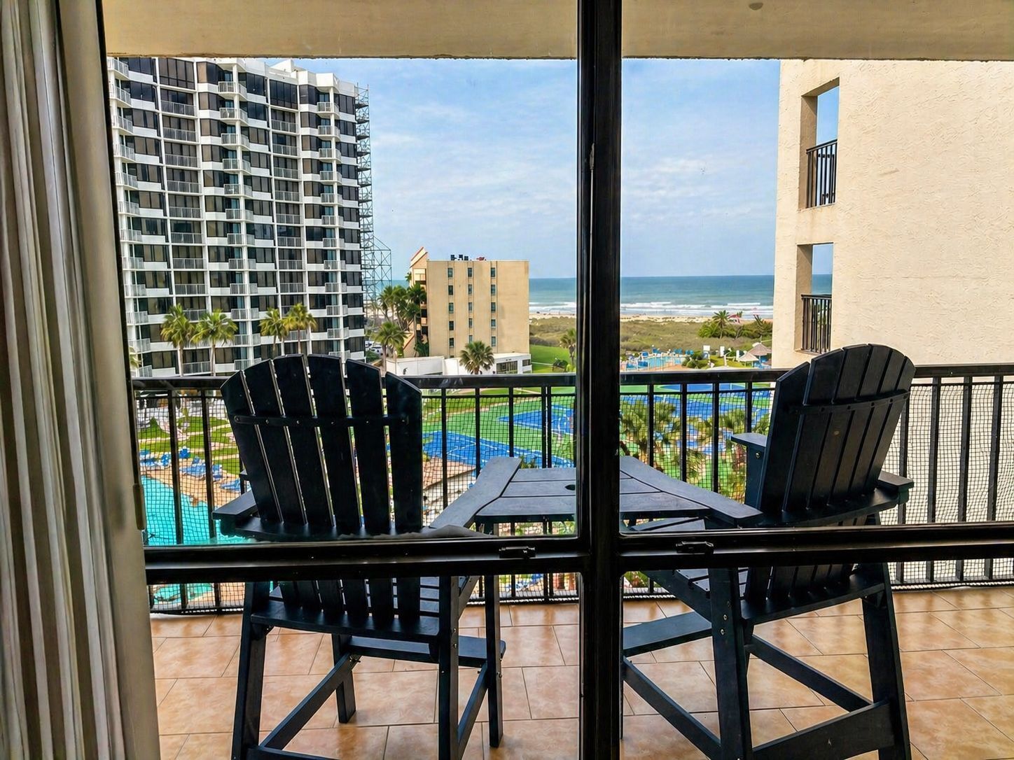 A balcony with two Adirondack chairs, a small table, and a view of a swimming pool, beach, and buildings.