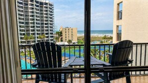 A balcony with two Adirondack chairs, a small table, and a view of a swimming pool, beach, and buildings. - High-Floor Saida Beachfront - Pool + Ocean View (South Padre Island)
