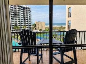 Ein Balkon mit zwei Adirondack-Stühlen, einem kleinen Tisch und Blick auf ein Schwimmbecken, einen Strand und Gebäude.