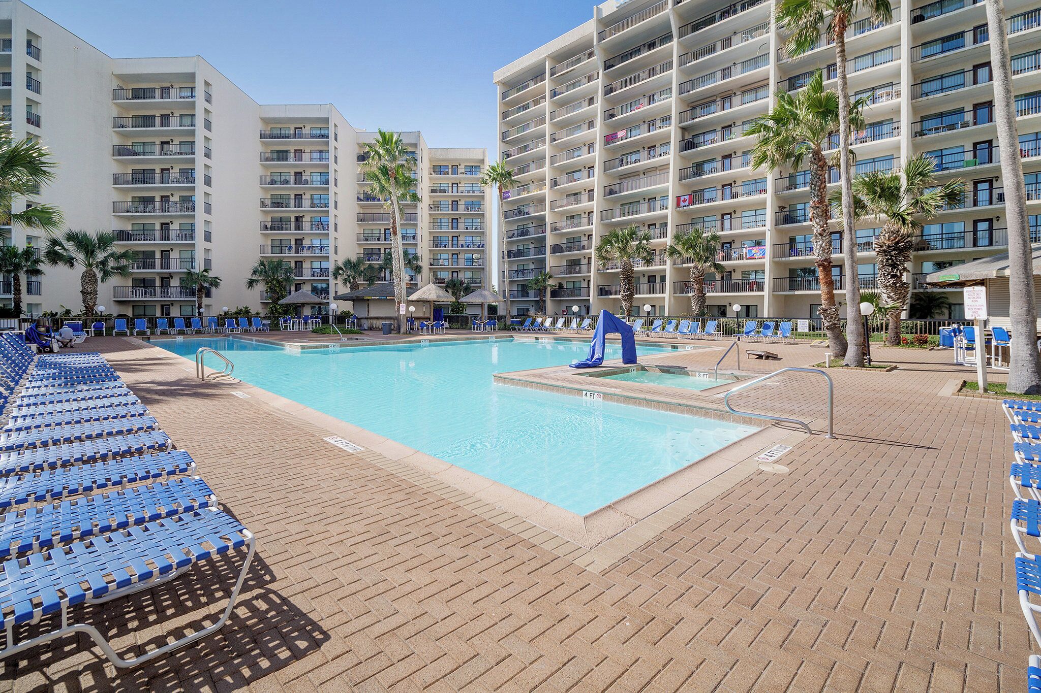 A hotel pool area with multiple levels of balconies, palm trees, and blue lounge chairs.