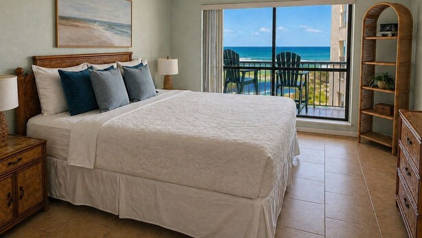 A bedroom with a bed, a ceiling fan, a painting, a nightstand, a lamp, a view of the ocean through a balcony door, and a wicker shelf. - High-Floor Saida Beachfront - Pool + Ocean View (South Padre Island)