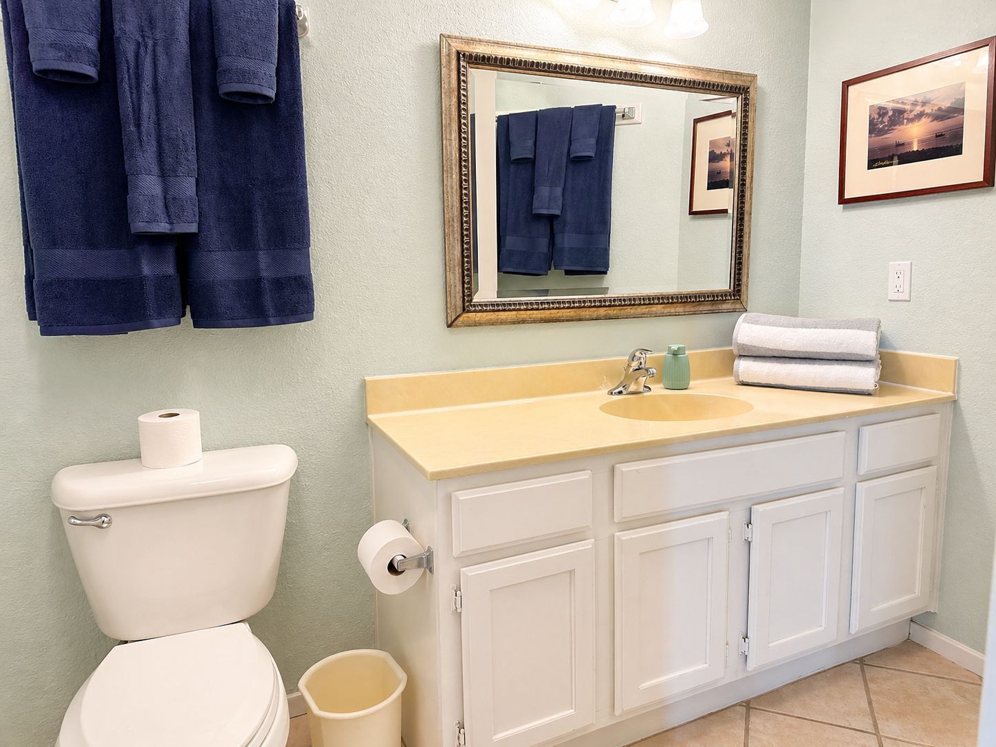 Une salle de bain équipée de toilettes, d’un lavabo, d’un miroir, de serviettes et d’un tableau accroché au mur.