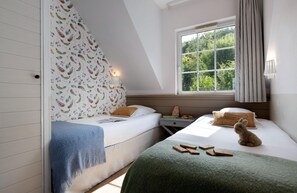 Un dormitorio con dos camas, una ventana con vista a follaje, una lámpara y un osito de peluche sobre una de las camas.