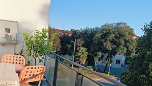 Uma varanda com mesa e cadeiras, corrimão e vista para árvores e edifícios.