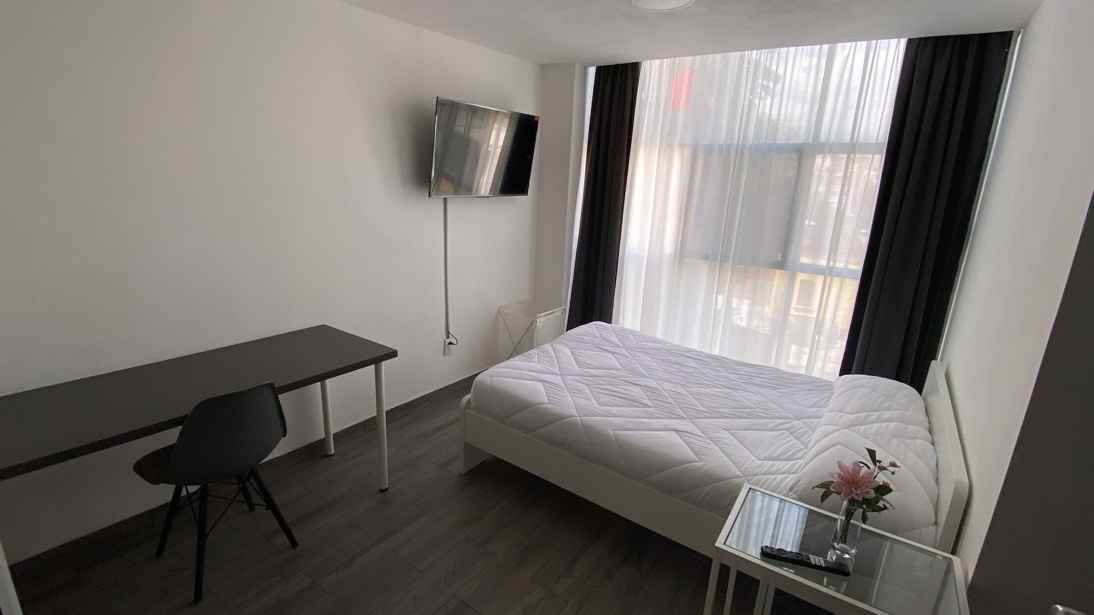 Een hotelkamer met een bed, een bureau, een stoel, een aan de muur bevestigde televisie en een raam met gordijnen.