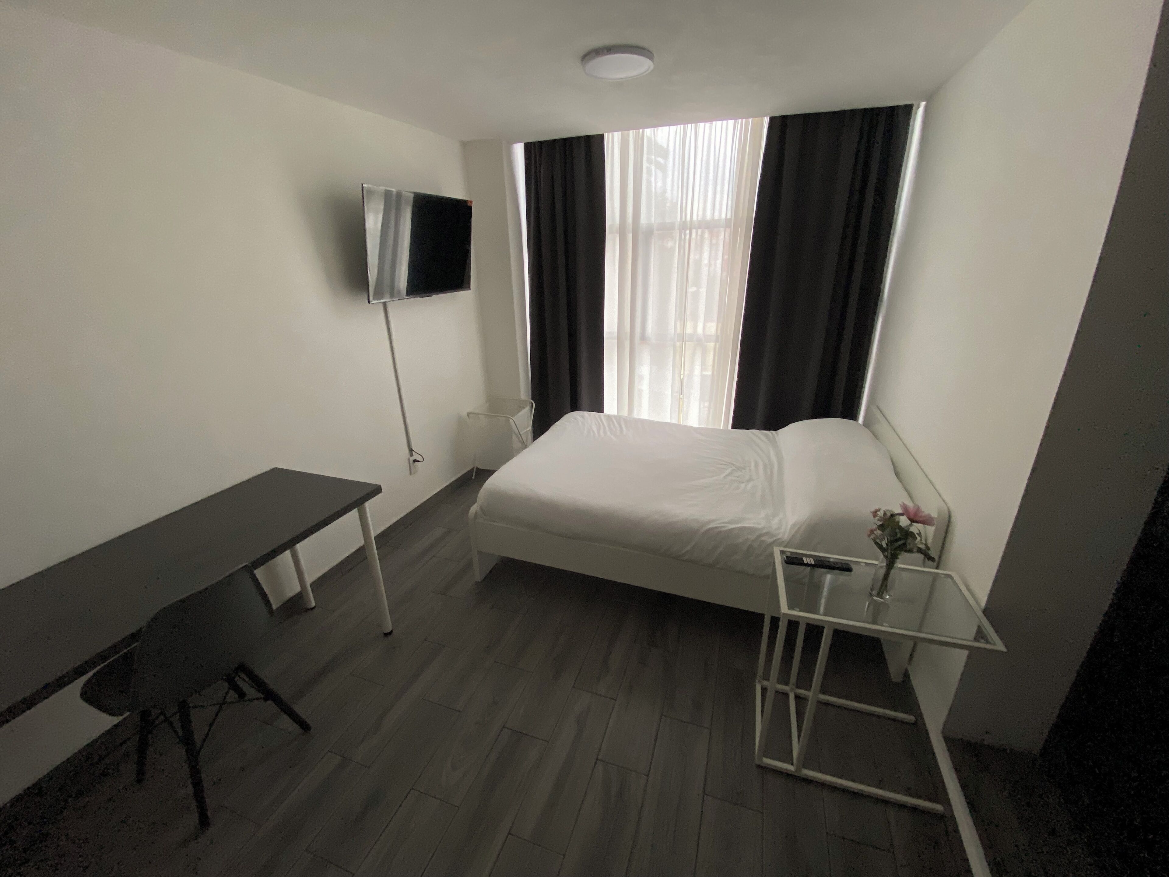 A small, modern hotel room with a bed, a desk, a TV, and a window with curtains.
