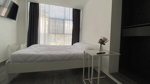 Ein modernes Hotelzimmer mit einem weißen Bett, einem kleinen Tisch mit einer Vase, einem Fernseher an der Wand und einem großen Fenster mit Vorhängen.