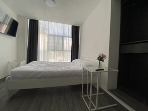 A modern hotel room with a white bed, a small table with a vase, a TV on the wall, and a large window with curtains. - Beautiful apartment near Angel de la Independencia (Cuauhtémoc)