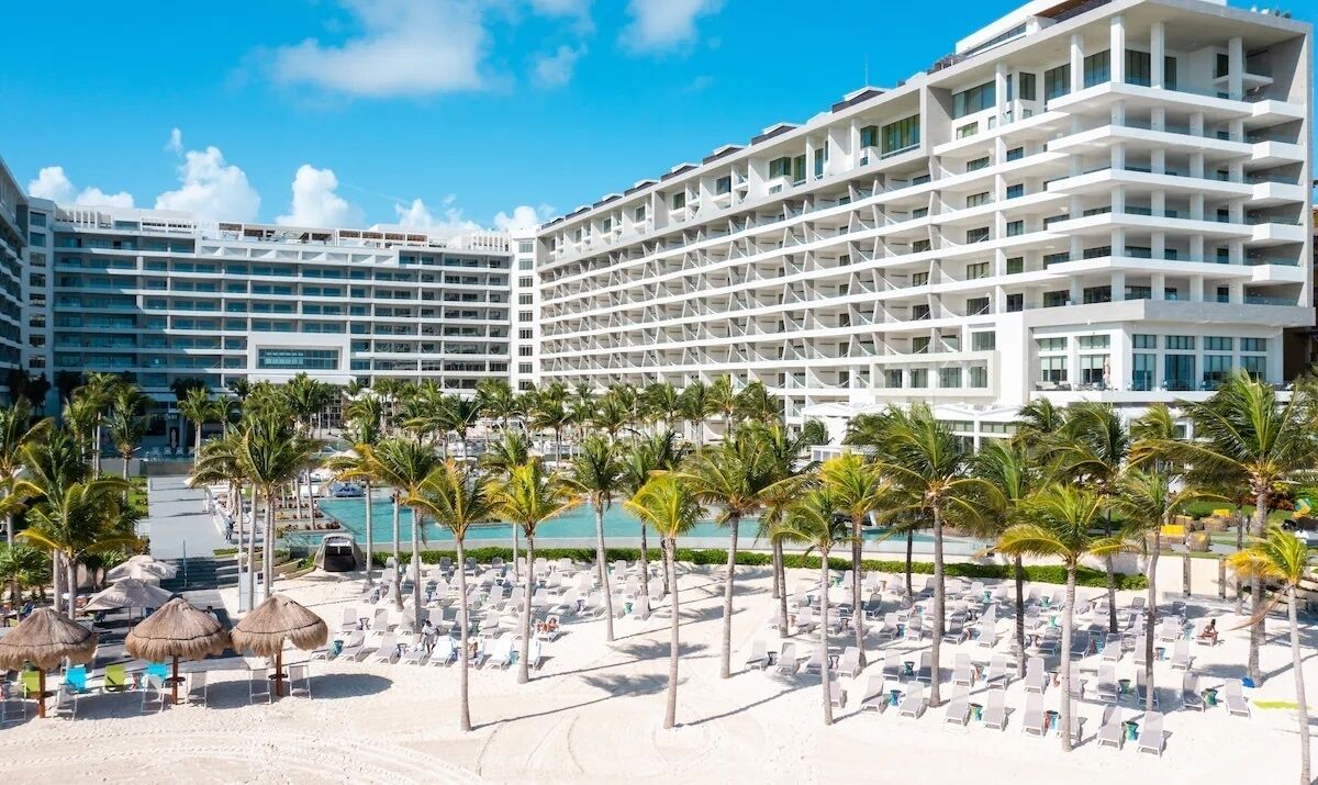 Een hotel met meerdere verdiepingen, een zwembad en een strandgedeelte met ligbedden en palmbomen.