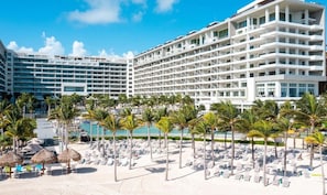 A hotel with multiple floors, a pool, and a beach area with sun loungers and palm trees.