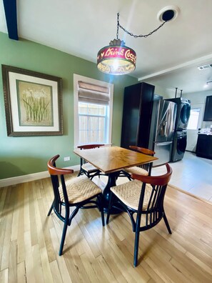 Un comedor con una mesa y sillas de madera, un cuadro en la pared, una ventana con persianas y una cocina al fondo.