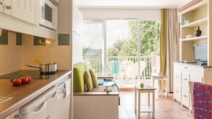 A kitchen with a microwave, stove, and sink; a dining area with a table and chairs; and a balcony with a view of greenery. - Family Apartment for 4 in Normandy (Branville)