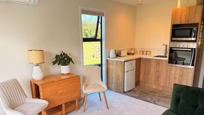 Eine kompakte Küche mit Mikrowelle, Backofen und Toaster, ein Holzesstisch mit Stühlen, eine Topfpflanze und Blick durch ein Fenster.