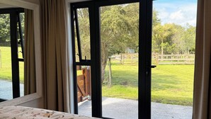 Ein Schlafzimmer mit einem großen Bett, Blick auf eine grüne Außenanlage durch eine Schiebetür und einem Holzstuhl in der Nähe der Tür.