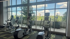 Et fitnesscenter med motionscykler og løbebånd, store vinduer samt udsigt til grønne områder og bygninger.
