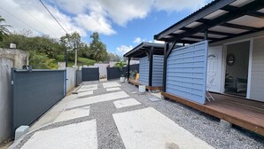 A covered outdoor area with a wooden deck, a grey fence, and a building with a sliding door. - Bungalow NANA - Comfort, calm and private terrace near CHU and airport (Les Abymes)