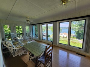 A covered porch with a table and chairs, large windows with a view of a lake and houses, and a ceiling fan.