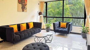 A modern living room with a sofa, armchair, and coffee table, large windows with curtains, and a painting on the wall. - Tranquil 2-bedroom Apartment With Nature and City Views (Nairobi)