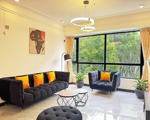 Ein modernes Wohnzimmer mit Sofa, Sessel und Couchtisch, großen Fenstern mit Vorhängen sowie einem Gemälde an der Wand.
