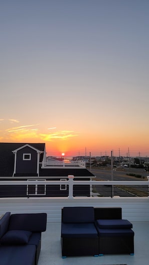 A balcony with two dark blue lounge chairs, a building with a white railing, and a sunset over a residential area.