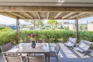 Une terrasse couverte avec une table et des chaises, entourée de verdure et offrant une vue sur des bâtiments au loin.