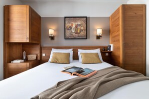 Habitación de hotel con una cama grande, cabecera de madera, dos lámparas de noche, un cuadro en la pared y una mesita de noche con lámpara y un libro.