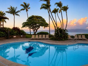 Ein Pool mit Liegestühlen, Palmen und Blick auf den Ozean bei Sonnenuntergang.
