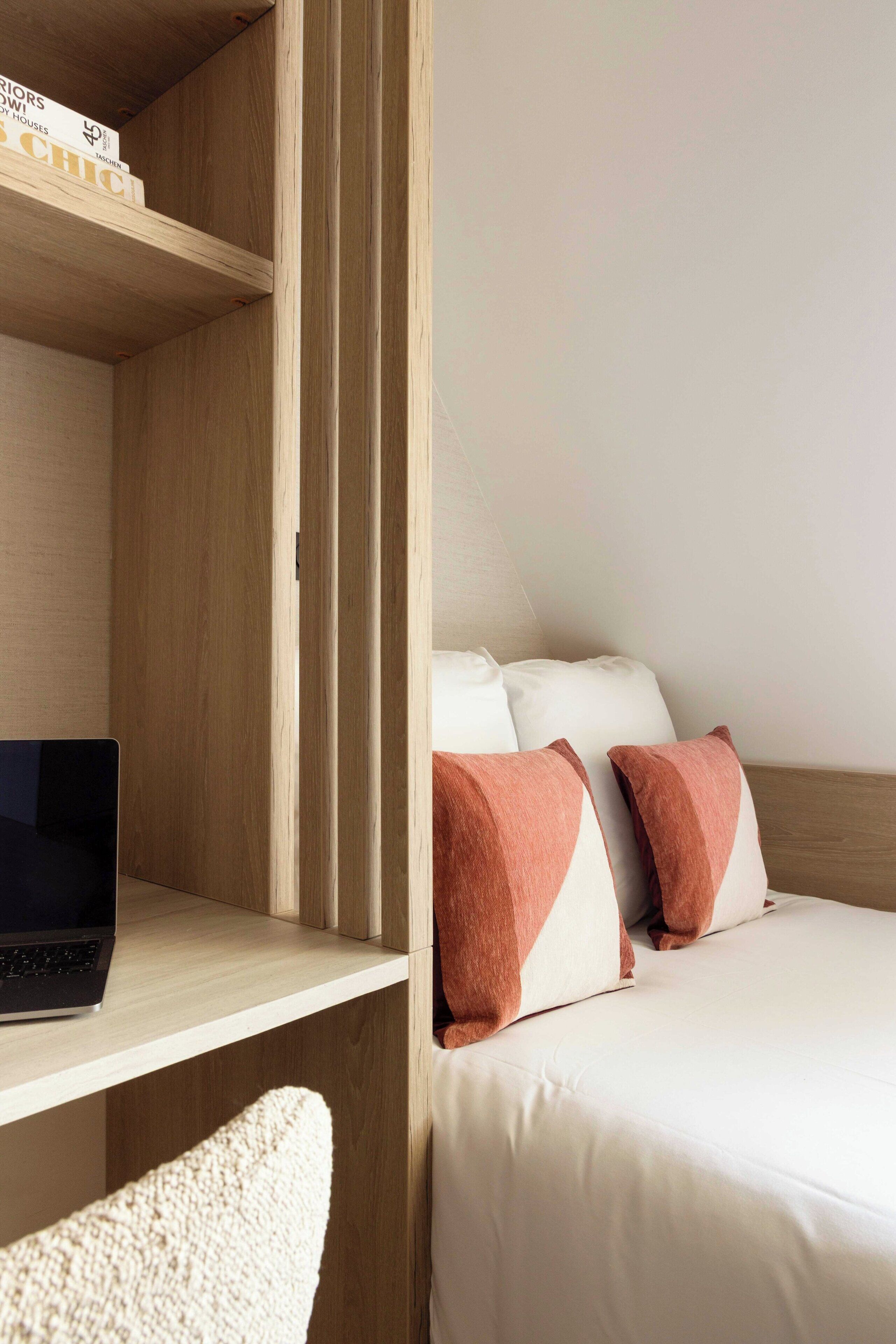 A hotel room with a wooden desk, a laptop, a bed with white and brown pillows, and a white chair.