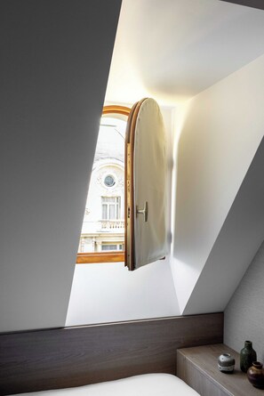 Ein Schlafzimmer mit schrägen Wänden, einem Fenster mit Blick auf ein Gebäude, einem Nachttisch mit einer Vase und einem Bett.