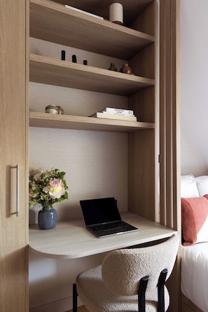 Ein Zimmer mit integriertem Schreibtisch, einem Laptop, einem Stuhl, einem Bücherregal und einem Bett mit Kissen.