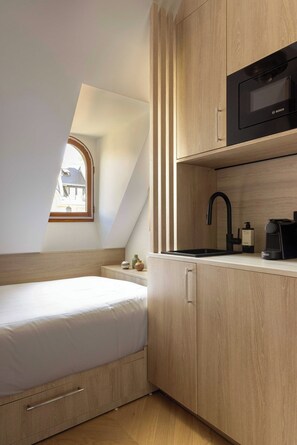 Ein kleines Schlafzimmer mit einem Einzelbett, einem Nachttisch, einer integrierten Mikrowelle und einem Fenster mit Blick auf ein Haus.