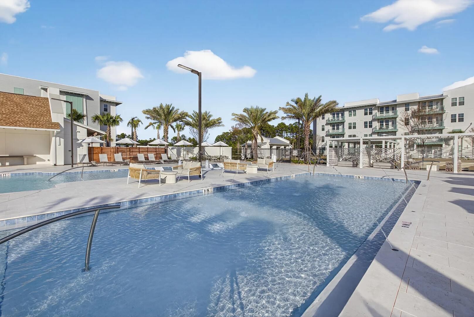 A swimming pool with lounge chairs and umbrellas, surrounded by palm trees and buildings.