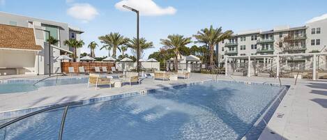 A swimming pool with lounge chairs and umbrellas, surrounded by palm trees and buildings.