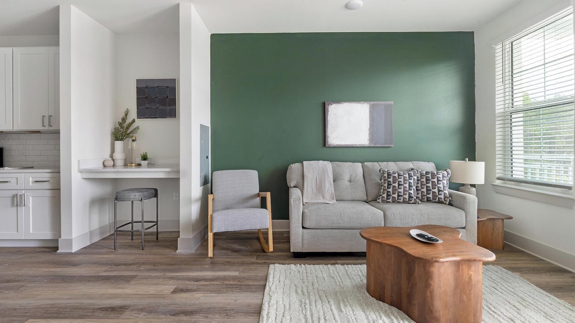 A modern living room with a green accent wall, a grey sofa, a wooden coffee table, and a kitchen area with white cabinets.