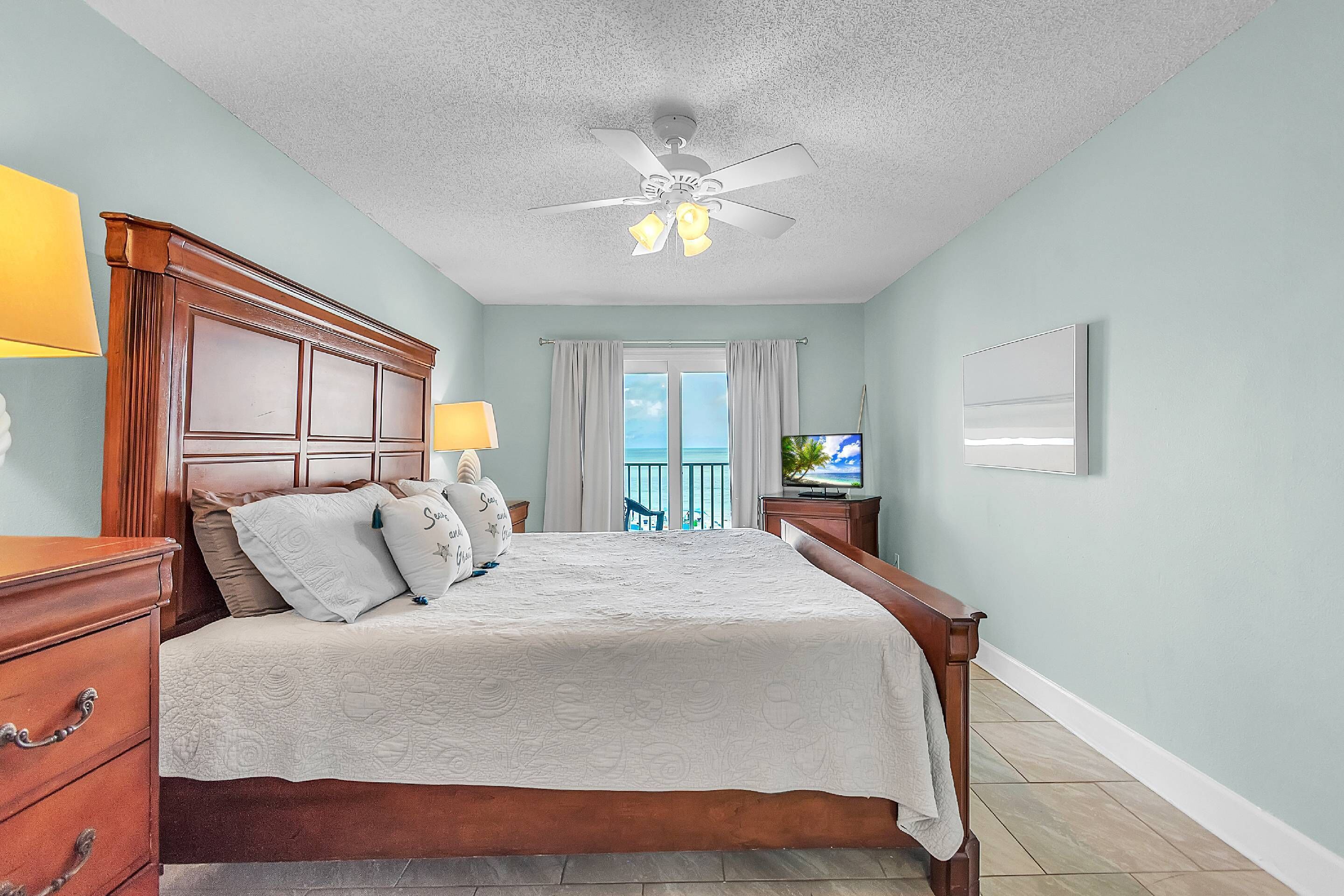 A bedroom with a wooden bed, a ceiling fan, a TV, and a view of the outdoors.