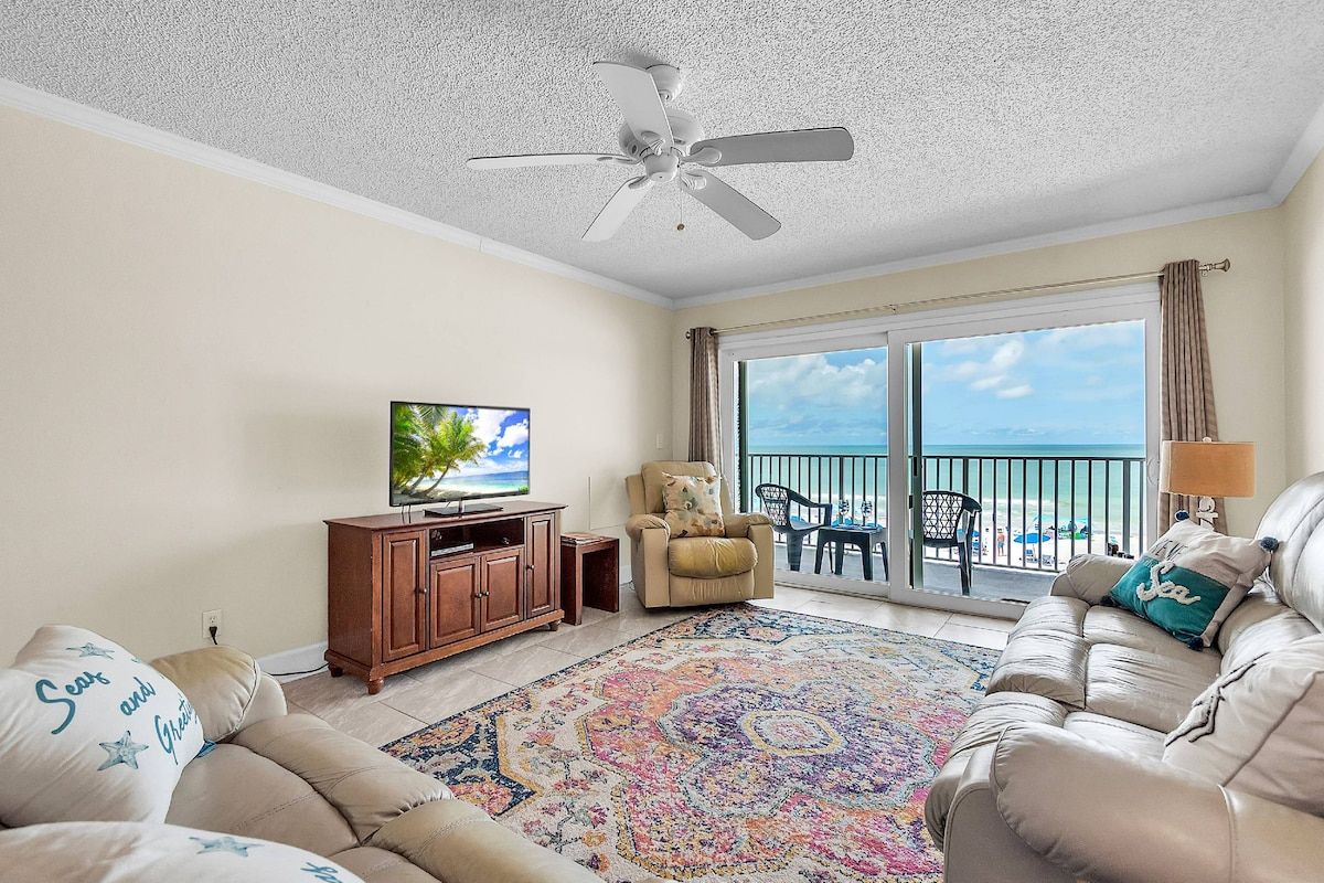 A living room with a ceiling fan, a television, a sofa, a chair, a patterned rug, and a balcony with a view of the ocean.